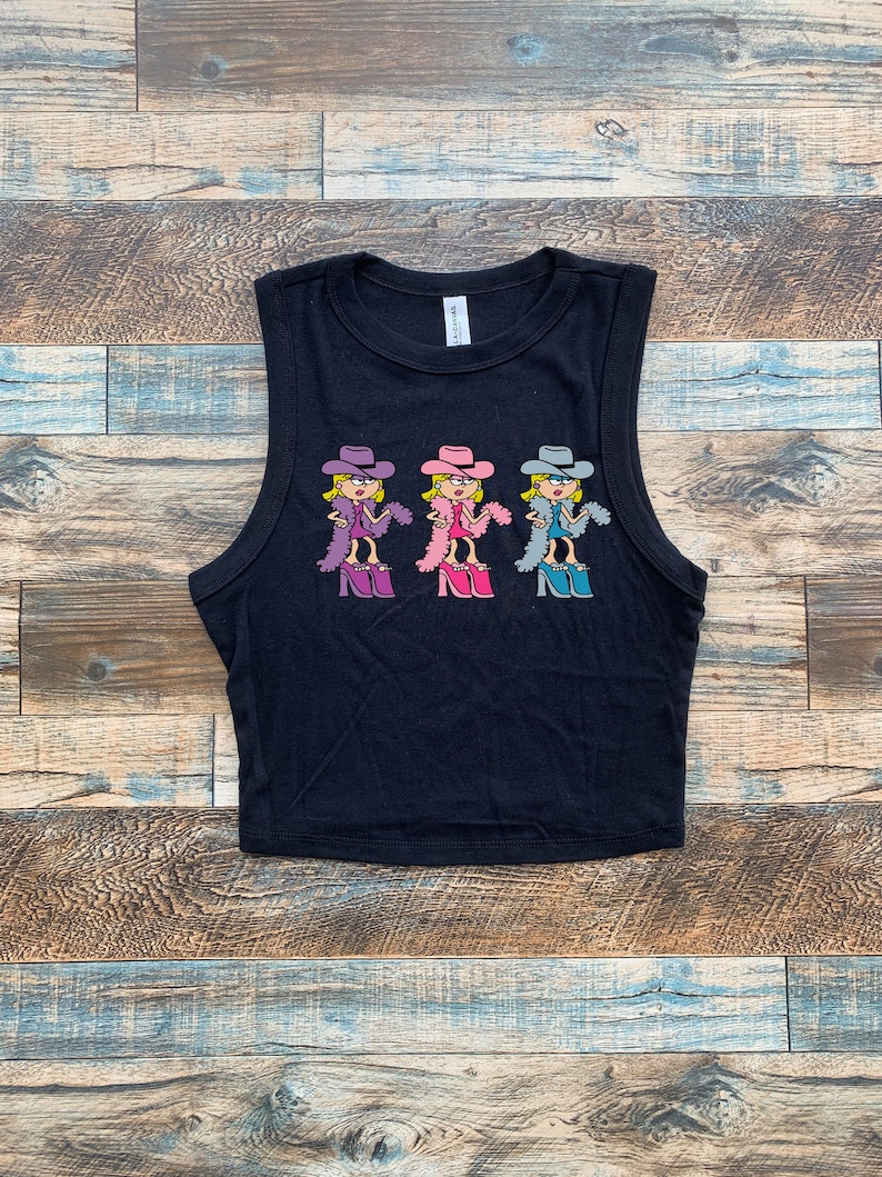 Op de afbeelding: Een zwarte mouwloze crop top met een grafische print van drie stripfiguren die cowboyhoeden, boa's en plateauschoenen dragen. De figuren zijn uitgevoerd in paarse, roze en blauwe kleurenschema's en horizontaal over de borst geplaatst.