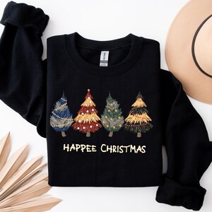 May include: Black sweatshirt featuring four embroidered Christmas trees in blue, red, green, and gold. The text "Happee Christmas" is printed in gold below the trees. A tan hat is visible in the background.