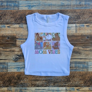 Puede incluir: Una camiseta corta azul claro con un gráfico colorido con personajes de dibujos animados en una cuadrícula. El texto "MOM VIBES" está impreso debajo en una fuente retro.