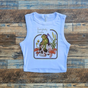 Könnte beinhalten: Ein hellblaues ärmelloses Crop-Top mit einem grafischen Aufdruck einer auf einem Schädel sitzenden Kröte, die eine Sense hält. Die Grafik ist von einem lila und gelben Rand umgeben. Der Text "Everybody Croaks" ist in Gelb über der Grafik gedruckt.