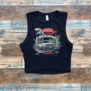 Può includere: Top corto nero con grafica di Jurassic Park. Il design include un dinosauro, un veicolo e la scritta "JURASSIC PARK" in rosso e bianco. Il top è senza maniche e ha uno scollo tondo.