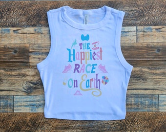 Può includere: Un top corto bianco senza maniche con il testo "The Happiest Race on Earth" in lettere colorate. Il testo è circondato da varie icone a tema Disney, tra cui le orecchie di Topolino, una tazza da tè, una scarpa da corsa e un castello.