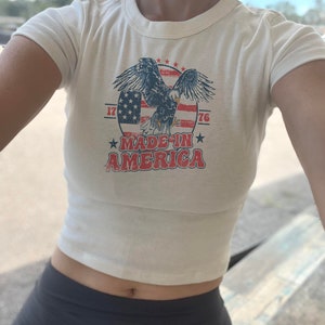 May include: White cropped t-shirt with a vintage-style graphic of an eagle flying over an American flag. The graphic also includes the text "Made in America" and the numbers "1776".