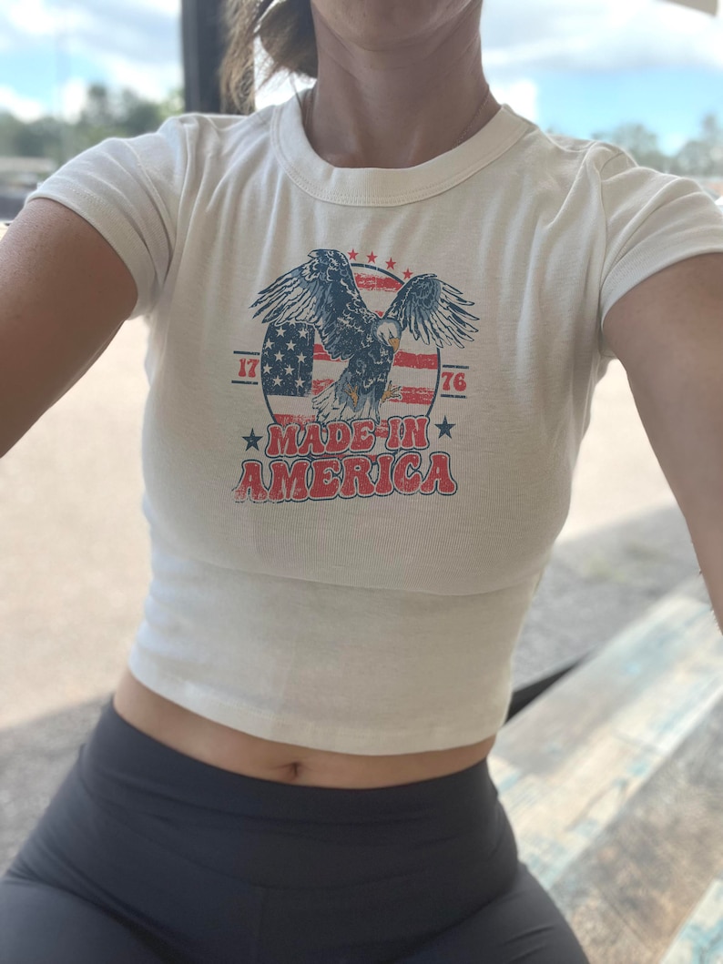 May include: White cropped t-shirt with a vintage-style graphic of an eagle flying over an American flag. The graphic also includes the text "Made in America" and the numbers "1776".
