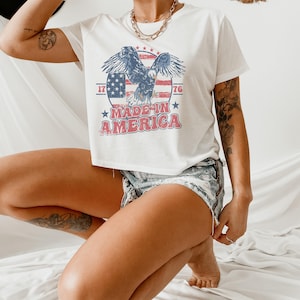 May include: White cropped t-shirt with a vintage-style graphic of an eagle flying over an American flag with the text "Made in America" and the numbers "1776".