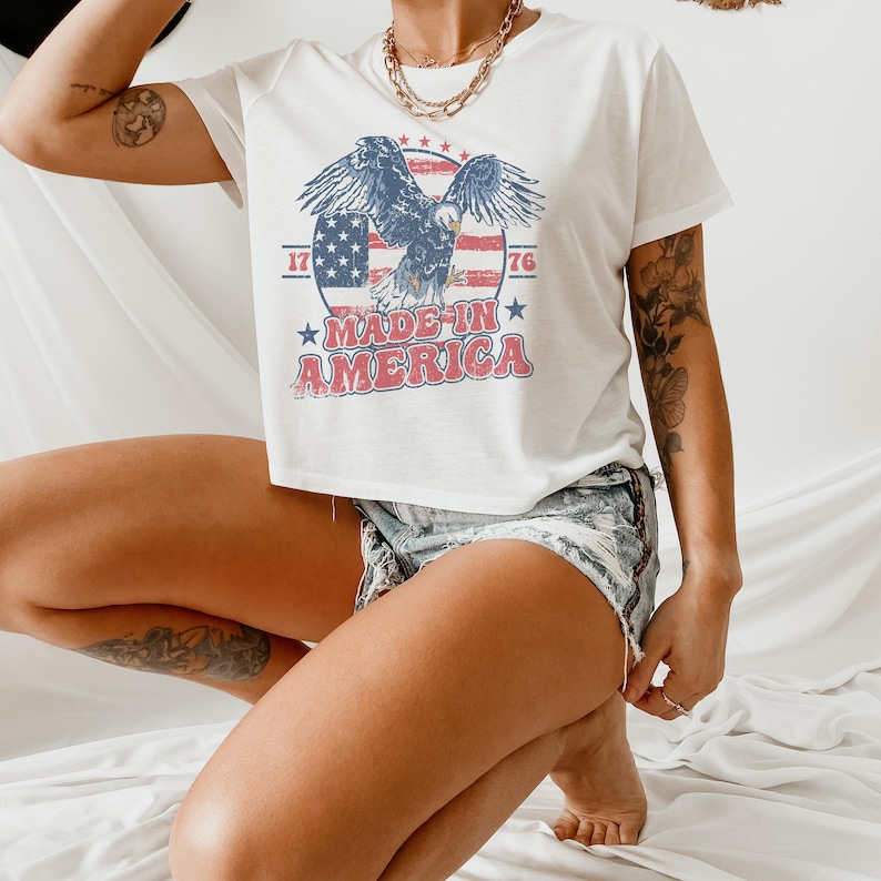 May include: White cropped t-shirt with a vintage-style graphic of an eagle flying over an American flag with the text "Made in America" and the numbers "1776".