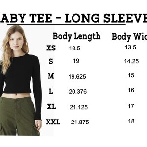 Puede incluir: Una camiseta de beb&eacute; negra de manga larga. La imagen incluye una tabla de tallas con medidas de largo y ancho del cuerpo en pulgadas para las tallas XS a XXL. La modelo lleva la camiseta negra con pantalones cargo verde oliva.