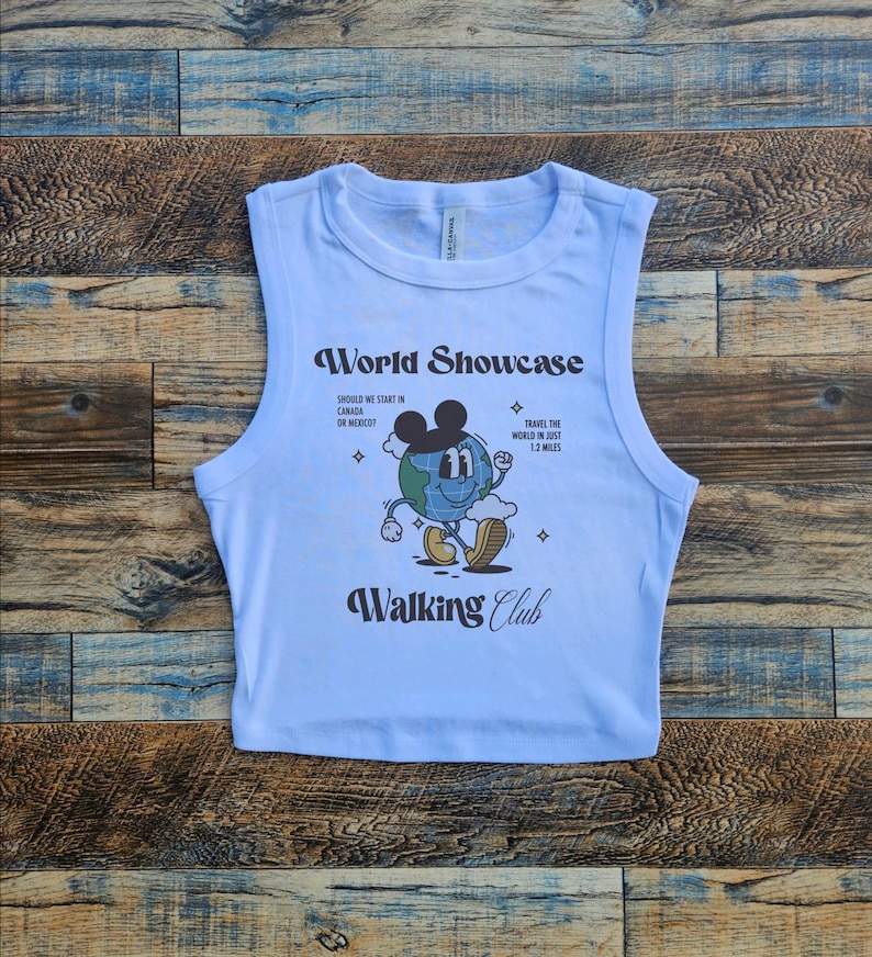 May include: Light blue sleeveless crop top with a graphic of a cartoon globe wearing Mickey Mouse ears and the text "World Showcase", "Should we start in Canada or Mexico?", "Travel the world in just 1.2 miles", and "Walking Club".