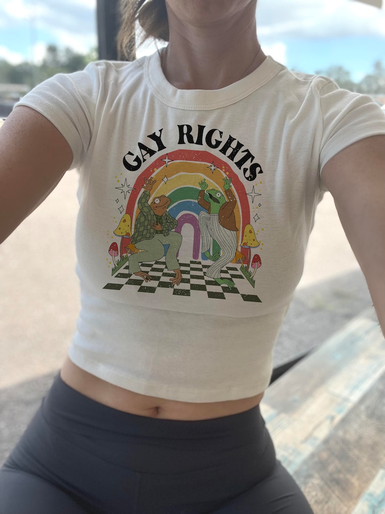 May include: Cream-colored crop top with the words "GAY RIGHTS" above a rainbow and two cartoon frogs dancing. The frogs are on a checkered floor with mushrooms. The top is short-sleeved.