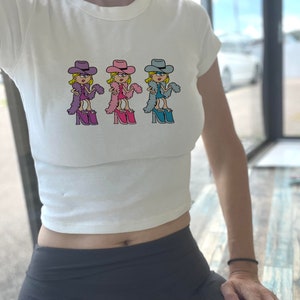 Op de afbeelding: Een wit kort T-shirt met korte mouwen en een herhalende grafische print van een stripfiguur met een cowboyhoed, een veren boa en plateauschoenen. Het personage is afgebeeld in drie verschillende kleurvariaties: paars, roze en blauw.