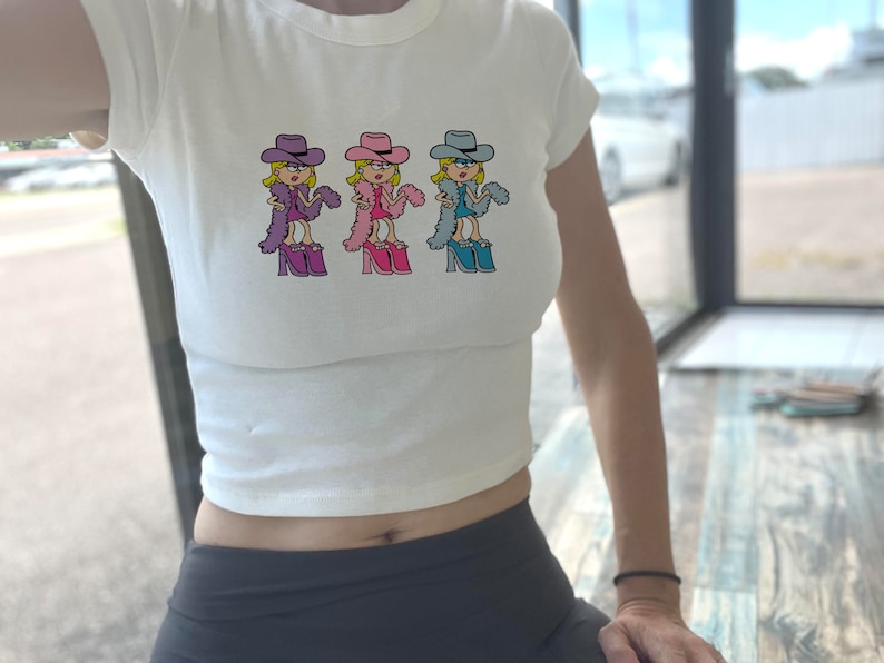 Op de afbeelding: Een wit kort T-shirt met korte mouwen en een herhalende grafische print van een stripfiguur met een cowboyhoed, een veren boa en plateauschoenen. Het personage is afgebeeld in drie verschillende kleurvariaties: paars, roze en blauw.