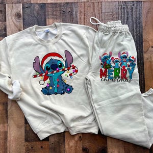 May include: A cream-coloured sweatshirt and sweatpants set featuring a cartoon Stitch character wearing a Santa hat and holding a candy cane. The sweatshirt has a Stitch design with Christmas lights. The sweatpants have a group of Stitch characters with the text "Merry Christmas".