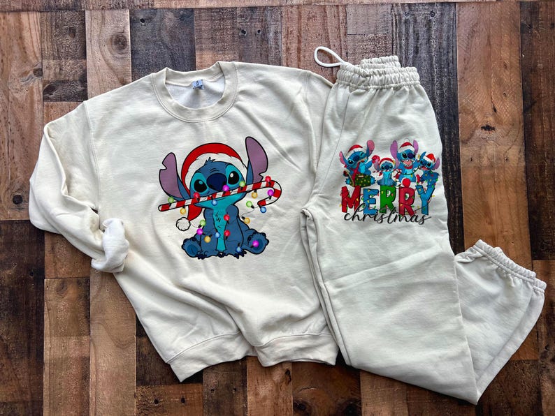 May include: A cream-coloured sweatshirt and sweatpants set featuring a cartoon Stitch character wearing a Santa hat and holding a candy cane. The sweatshirt has a Stitch design with Christmas lights. The sweatpants have a group of Stitch characters with the text "Merry Christmas".