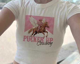 Puede incluir: Camiseta blanca recortada con un gráfico rosa de un vaquero montando un caballo con alas. El gráfico tiene el texto "Pucker Up Cowboy" debajo.