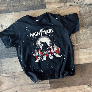 Puede incluir: Camiseta negra con salpicaduras de pintura blanca y un gr&aacute;fico de "Pesadilla antes de Navidad". El gr&aacute;fico presenta a Jack Skellington, Sally y otros personajes en un paso de peatones frente a una luna llena.