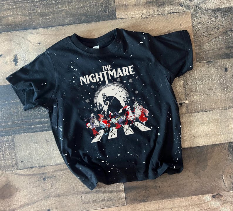 Puede incluir: Camiseta negra con salpicaduras de pintura blanca y un gr&aacute;fico de "Pesadilla antes de Navidad". El gr&aacute;fico presenta a Jack Skellington, Sally y otros personajes en un paso de peatones frente a una luna llena.