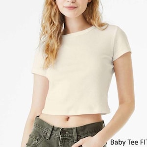 Op de afbeelding: Een cr&egrave;mekleurig baby-T-shirt met korte mouwen, een ronde halslijn en een kortere zoom. Het shirt wordt gedragen met een donker olijfgroene spijkerbroek. De tekst "Baby Tee FIT" staat in de rechterbenedenhoek van de afbeelding.