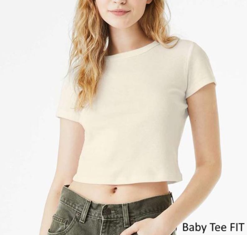 Op de afbeelding: Een cr&egrave;mekleurig baby-T-shirt met korte mouwen, een ronde halslijn en een kortere zoom. Het shirt wordt gedragen met een donker olijfgroene spijkerbroek. De tekst "Baby Tee FIT" staat in de rechterbenedenhoek van de afbeelding.