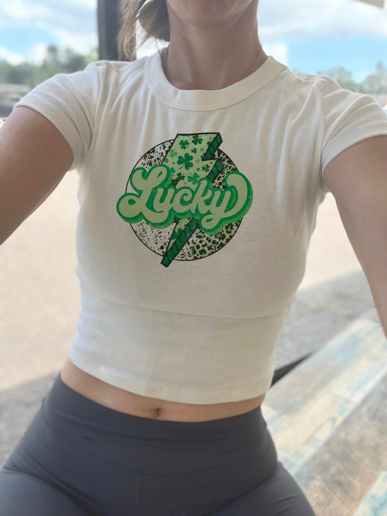 May include: White crop top with a green and black graphic of a lightning bolt with the word "Lucky" in a retro font. The graphic is surrounded by shamrocks.