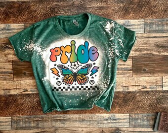 Puede incluir: Camiseta verde con un diseño salpicado de lejía que presenta la palabra "pride" en colores arcoíris y una mariposa con alas arcoíris. El diseño también incluye un signo de paz, un rayo y un patrón a cuadros.