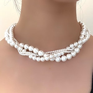 Könnte beinhalten: Eine mehrreihige Perlenkette mit einem gedrehten Design. Die Kette besteht aus weißen Perlen und wird von einer Person getragen.