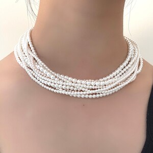 May include: A white pearl choker necklace with multiple strands of pearls. The necklace is worn by a person with a black top.