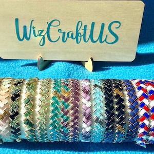 May include: A collection of twelve woven bead bracelets displayed on a cylindrical holder, featuring various color patterns including metallic, green, purple, blue, and a red, white, and blue design. A wooden sign behind reads WizCraftUS.