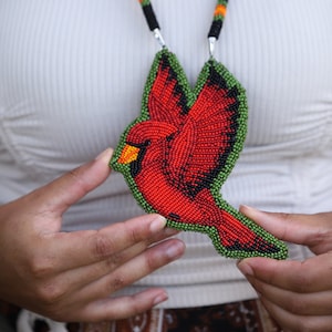 以下が含まれることがあります： 赤いカージナルのペンダントが付いたビーズネックレス。カージナルは羽を広げて飛んでいます。ネックレスは黒、赤、緑のビーズで作られています。