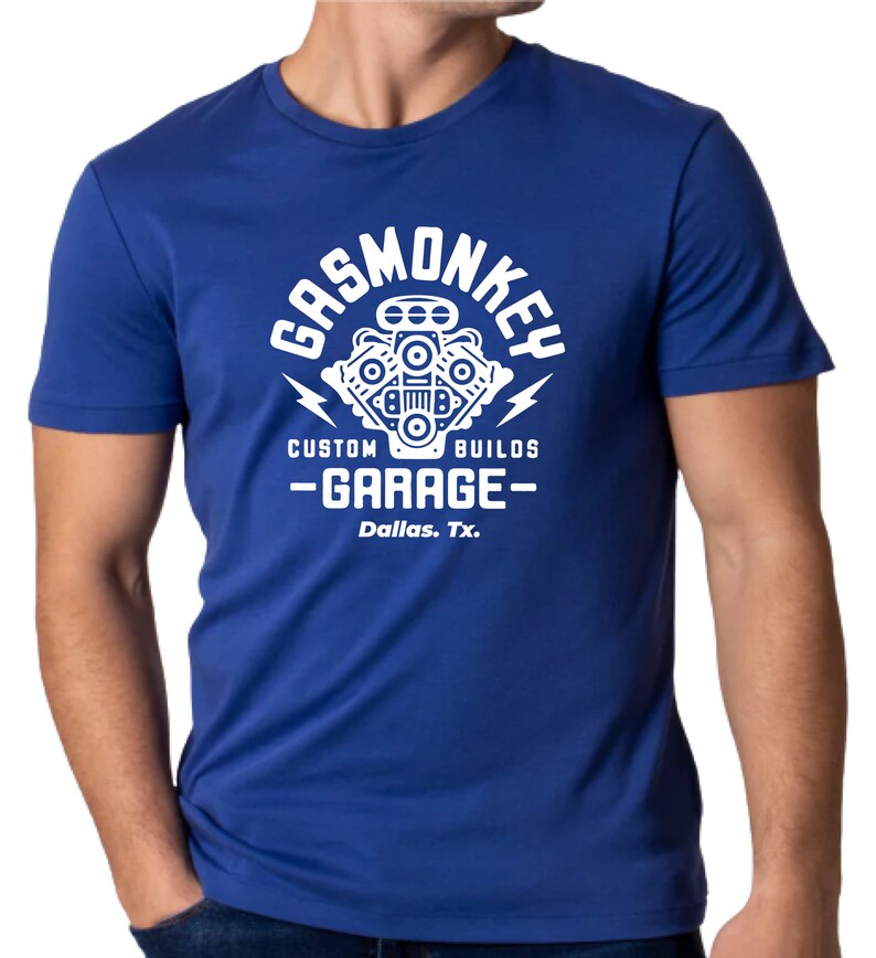 Pode incluir: Uma camiseta azul royal com um gr&aacute;fico branco de um motor estilizado com o texto "Gas Monkey Custom Builds - Garage - Dallas, Tx."