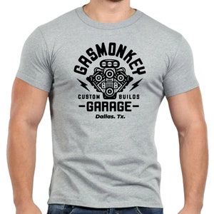 Pode incluir: Uma camiseta cinza com um gr&aacute;fico preto de um motor estilizado com o texto "Gas Monkey Custom Builds - Garage - Dallas, Tx."