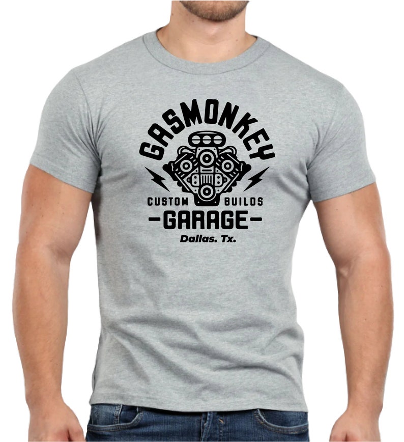 Pode incluir: Uma camiseta cinza com um gr&aacute;fico preto de um motor estilizado com o texto "Gas Monkey Custom Builds - Garage - Dallas, Tx."
