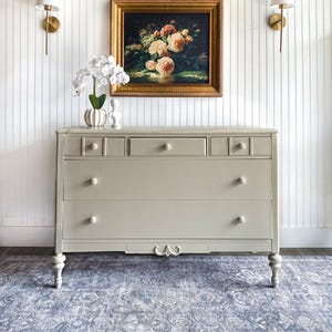 May include: A light gray wooden dresser featuring three large drawers and three smaller top drawers with round knobs. The piece stands on four turned legs and includes a decorative carved detail at the center of the bottom apron.