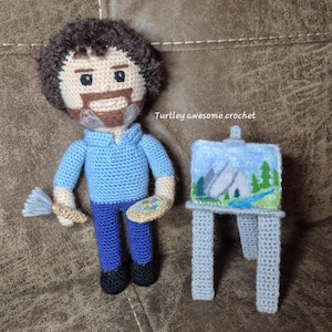 May include: Crocheted figure of a person with brown hair and a beard holding a paintbrush and a palette. The figure is wearing a blue shirt and blue pants. The figure is standing next to a crocheted easel with a painting of a mountain scene.