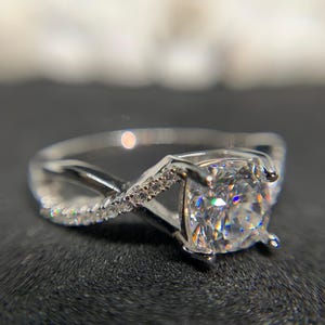 Può includere: Un anello d'argento con una grande pietra preziosa quadrata trasparente incastonata al centro. La fascia è attorcigliata e presenta pietre preziose più piccole sui lati.