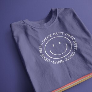 K&ouml;nnte beinhalten: Lilafarbenes T-Shirt mit einem wei&szlig;en Smiley und dem Text "Choose Happy. Choose Happy. Choose Happy. Choose Happy."