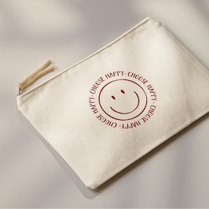 K&ouml;nnte beinhalten: Eine wei&szlig;e Canvas-Rei&szlig;verschlusstasche mit einem roten Smiley und dem Text "Choose Happy. Choose Happy. Choose Happy. Choose Happy." rundherum gedruckt.