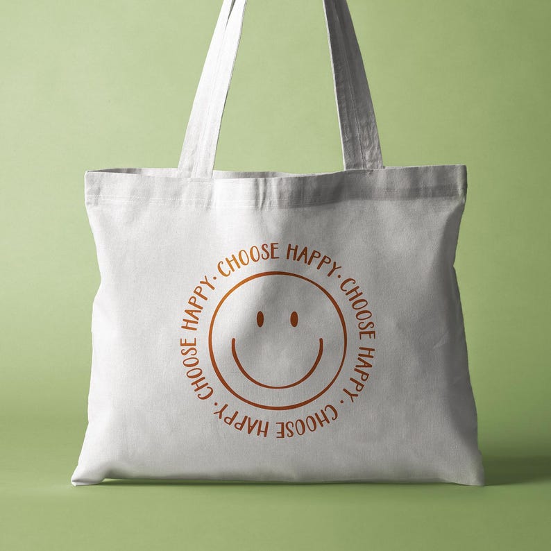 K&ouml;nnte beinhalten: Wei&szlig;e Canvas-Tragetasche mit einem braunen Smiley und dem Text "Choose Happy. Choose Happy. Choose Happy. Choose Happy." in einem Kreis um den Smiley gedruckt.