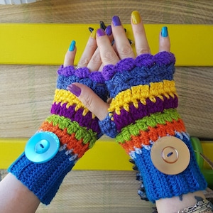 Könnte beinhalten: Ein Paar handgefertigte, farbenfrohe Fingerhandschuhe. Die Handschuhe sind mit blauer, lila, gelber, orangefarbener und grüner Wolle gehäkelt. Jeder Handschuh hat einen großen Knopf am Handgelenk. Der Hintergrund ist eine gelbe Oberfläche.