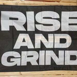 Puede incluir: Bandera negra y blanca con el texto "RISE AND GRIND" en letras blancas grandes y en negrita.