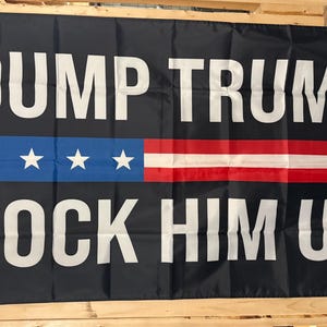 Pode incluir: Uma bandeira azul escura com texto branco que diz "DUMP TRUMP" e "LOCK HIM UP". A bandeira apresenta um design que se assemelha à bandeira americana, com uma faixa azul e estrelas brancas, e listras vermelhas e brancas.