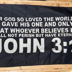 May include: Black flag with white text. The text reads: "FOR GOD SO LOVED THE WORLD THAT HE GAVE HIS ONE AND ONLY SON, THAT WHOEVER BELIEVES IN HIM SHALL NOT PERISH BUT HAVE ETERNAL LIFE. JOHN 3:16."