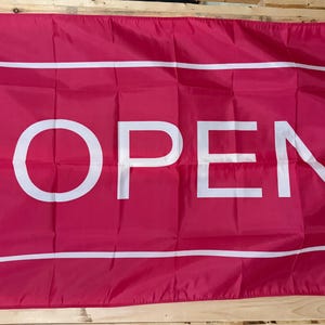 May include: A rectangular flag in a vibrant magenta pink color featuring the word OPEN printed in large, bold, white capital letters. The text is enclosed within a thin white rectangular border, and the flag includes a white side strip with grommets.