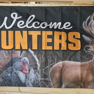 May include: A black welcome banner with the text "Welcome Hunters" in white and orange lettering. The banner features a realistic image of a brown deer with large antlers and a wild turkey.
