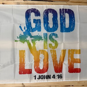 May include: White flag with the words "GOD IS LOVE" in large, colorful letters. The text is in a distressed font, with a rainbow-colored tree graphic. The Bible verse "1 JOHN 4:16" is printed at the bottom.