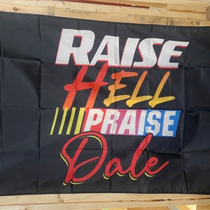 Könnte beinhalten: Schwarze Flagge mit dem Text "RAISE HELL PRAISE DALE" in bunten, stilisierten Buchstaben.