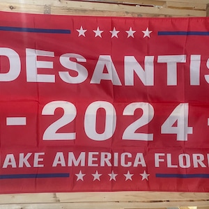 May include: A red political campaign flag with the text "DESANTIS - 2024 - MAKE AMERICA FLORIDA" in white lettering. The flag has a blue border and five white stars in the top right corner and five white stars in the bottom right corner.