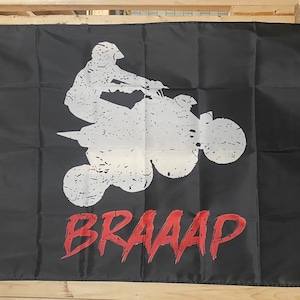 Puede incluir: Bandera negra con una silueta blanca de una persona conduciendo un ATV. El texto "BRAAAP" está escrito en rojo debajo del ATV.