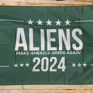 May include: A green flag with white text that reads "ALIENS MAKE AMERICA GREEN AGAIN 2024" with five white stars above the word "ALIENS" and three white stars on each side of the year "2024".