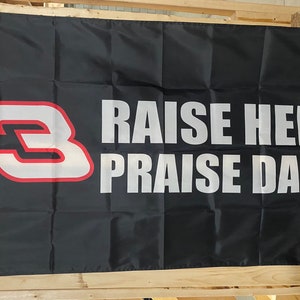 Könnte beinhalten: Schwarze Flagge mit einer weißen Nummer "3" mit rotem Rand und dem Text "RAISE HELL PRAISE DALE" in weiß.