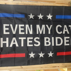 May include: A black flag with a red, white and blue border. The flag has three white stars on each side of the border and the text "EVEN MY CAT HATES BIDEN" in white letters.